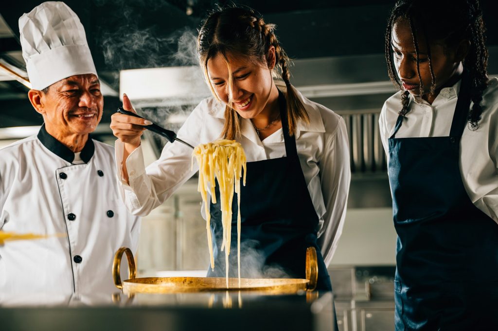 Chef educates in kitchen. Schoolgirls make Japanese noodle. Kids and teacher at stove. Smiling